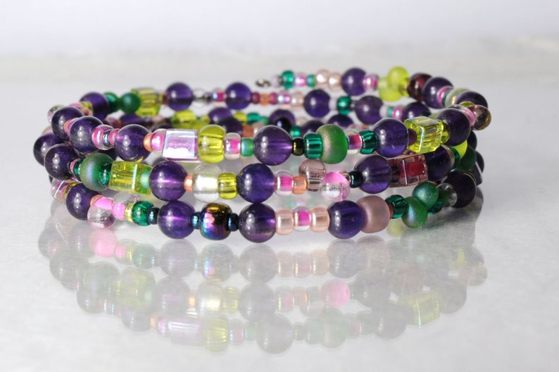 Op de afbeelding: Een meerstrengige armband met paarse, groene, roze en gele kralen. De armband is gemaakt van verschillende maten en vormen kralen, waaronder ronde, vierkante en rechthoekige.