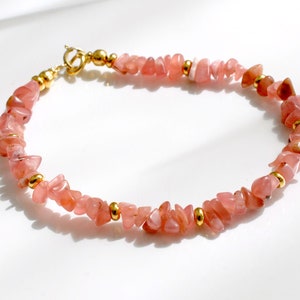 Könnte beinhalten: Ein zartes Armband aus rosafarbenen Edelsteinen mit goldenen Akzenten. Das Armband besteht aus kleinen, unregelmäßig geformten Steinen und hat einen goldenen Verschluss.