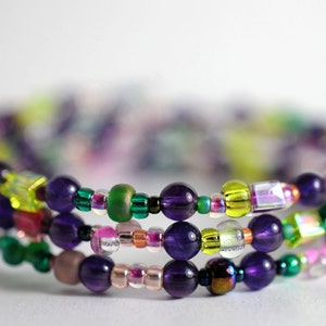 Op de afbeelding: Een meerstrengige armband met paarse, groene, gele en roze kralen. De armband is gemaakt van verschillende materialen, waaronder glas, metaal en edelstenen.