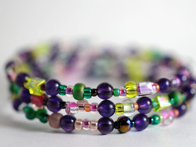 Op de afbeelding: Een meerstrengige armband met paarse, groene, gele en roze kralen. De armband is gemaakt van verschillende materialen, waaronder glas, metaal en edelstenen.