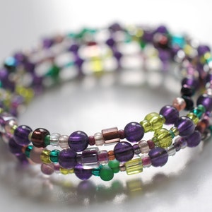 Op de afbeelding: Een meerstrengige armband met paarse, groene en bruine kralen. De armband is gemaakt van kleine, ronde kralen en is meerdere keren om de pols gewikkeld.