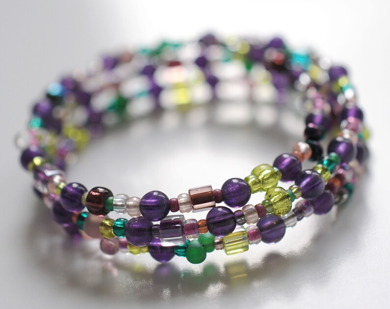 Op de afbeelding: Een meerstrengige armband met paarse, groene en bruine kralen. De armband is gemaakt van kleine, ronde kralen en is meerdere keren om de pols gewikkeld.
