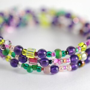 Op de afbeelding: Een meerstrengige armband met paarse, groene, roze en gele kralen. De armband is gemaakt van kralen in verschillende maten en vormen, waaronder ronde, vierkante en ovale.