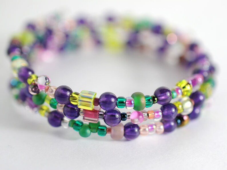 Op de afbeelding: Een meerstrengige armband met paarse, groene, roze en gele kralen. De armband is gemaakt van kralen in verschillende maten en vormen, waaronder ronde, vierkante en ovale.