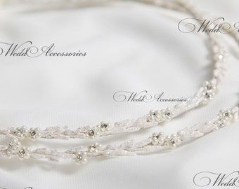 May include: Two white, beaded, and sparkly wedding crowns, or stefana, are intertwined and laid out on a white fabric background. The crowns are decorated with small white pearls and clear rhinestones.
