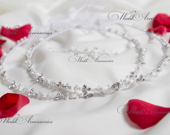May include: Two white and silver wedding crowns with pearl and crystal accents. The crowns are on a white surface with red rose petals.