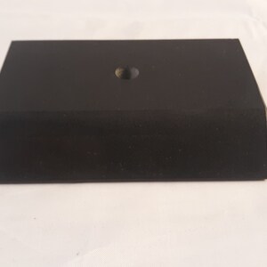 May include: A rectangular black display base with a matte finish and a single centered circular hole on the top surface. The object features beveled edges along the front and sides, creating a low-profile, solid block structure for mounting items.