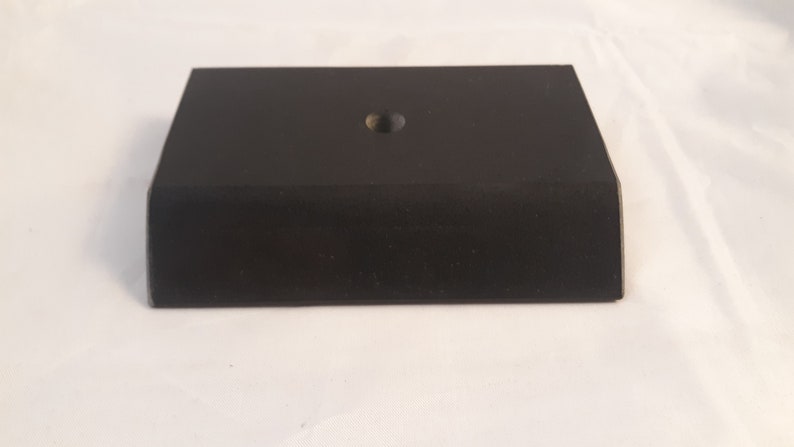 May include: A rectangular black display base with a matte finish and a single centered circular hole on the top surface. The object features beveled edges along the front and sides, creating a low-profile, solid block structure for mounting items.