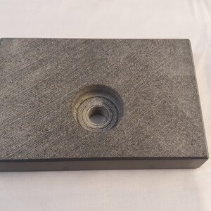May include: A rectangular, dark gray block with a textured, stone-like surface. The object features a centered, circular recessed area that contains a smaller, deep hole in the middle, showing concentric ring patterns from the manufacturing process.