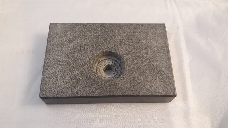 May include: A rectangular, dark gray block with a textured, stone-like surface. The object features a centered, circular recessed area that contains a smaller, deep hole in the middle, showing concentric ring patterns from the manufacturing process.