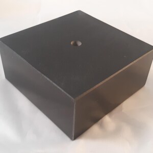 May include: A black square pedestal with a small hole in the center. The pedestal is made of wood and has a smooth finish.