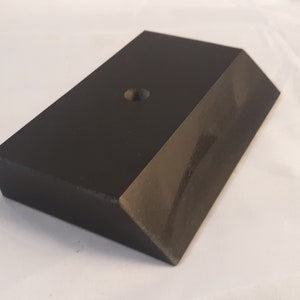 May include: A rectangular black block with a smooth, matte finish and a beveled edge along one side. The top surface features a single, centered circular hole, creating a simple geometric form with clean lines and a solid, uniform dark appearance.