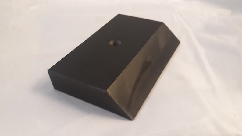 May include: A rectangular black block with a smooth, matte finish and a beveled edge along one side. The top surface features a single, centered circular hole, creating a simple geometric form with clean lines and a solid, uniform dark appearance.