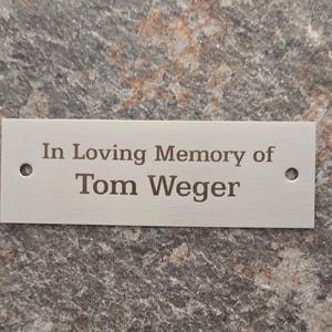 Könnte beinhalten: Eine rechteckige Gedenktafel aus silberfarbenem Metall mit der eingravierten Aufschrift In Loving Memory of Tom Weger. Die Tafel hat eine glatte, gebürstete Oberfläche und verfügt über zwei kleine kreisförmige Befestigungslöcher.