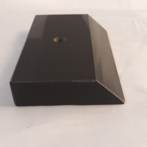 May include: A rectangular black block with a smooth, reflective surface and a beveled edge on one side. The top surface features a single, centered circular hole. The object has a solid, uniform dark appearance with sharp, clean geometric lines.