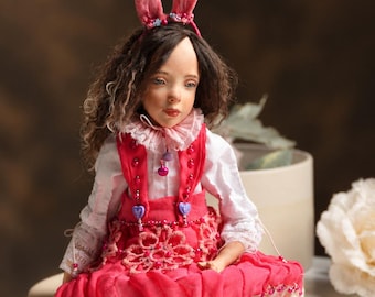 May include: A doll with dark wavy hair wearing pink velvet bunny ears, a white collared shirt, and a bright pink pinafore dress with lace trim, bead embellishments, and heart-shaped buttons. The doll wears dark leggings and light pink shoes.