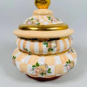May include: A decorative ceramic jar with a lid. The jar is painted with a peach and white checkered pattern, accented with gold trim and floral designs. The lid is topped with a gold sphere.