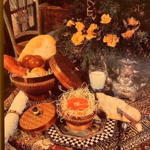K&ouml;nnte beinhalten: Ein Stillleben mit Brot, Br&ouml;tchen und einer Grapefruit-H&auml;lfte in Weidenk&ouml;rben. Ein Glas Milch und eine silberne Kanne stehen auf einer gemusterten Tischdecke. Gelbe Rosen und Trauben schaffen eine rustikale Atmosph&auml;re.