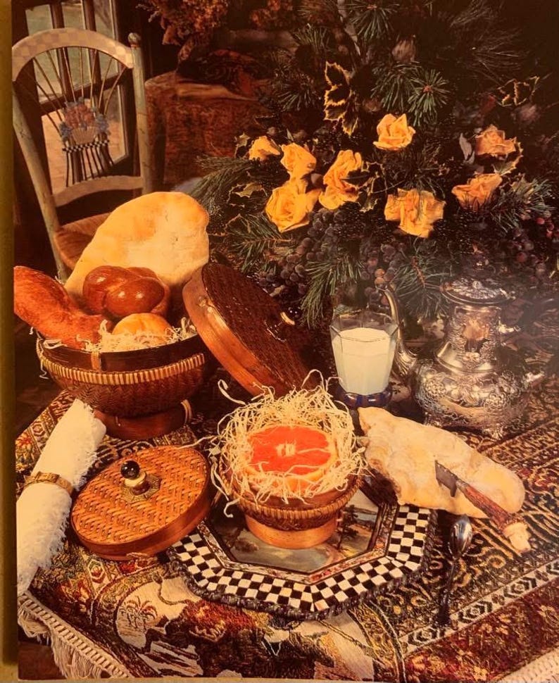 K&ouml;nnte beinhalten: Ein Stillleben mit Brot, Br&ouml;tchen und einer Grapefruit-H&auml;lfte in Weidenk&ouml;rben. Ein Glas Milch und eine silberne Kanne stehen auf einer gemusterten Tischdecke. Gelbe Rosen und Trauben schaffen eine rustikale Atmosph&auml;re.