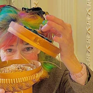 K&ouml;nnte beinhalten: Eine Frau mit Regenbogenhaar h&auml;lt einen kleinen, gewebten Korb mit einer klaren Glasschale mit einem Backwaren. Sie h&auml;lt auch eine kleine Holzkiste mit einem gr&uuml;nen und blauen Design. Der Hintergrund umfasst ein Gem&auml;lde und dekorative Verzierungen.