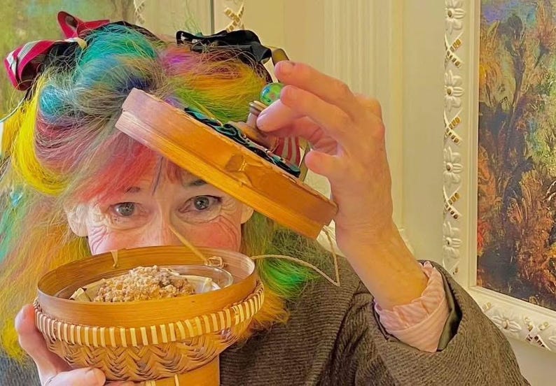 K&ouml;nnte beinhalten: Eine Frau mit Regenbogenhaar h&auml;lt einen kleinen, gewebten Korb mit einer klaren Glasschale mit einem Backwaren. Sie h&auml;lt auch eine kleine Holzkiste mit einem gr&uuml;nen und blauen Design. Der Hintergrund umfasst ein Gem&auml;lde und dekorative Verzierungen.