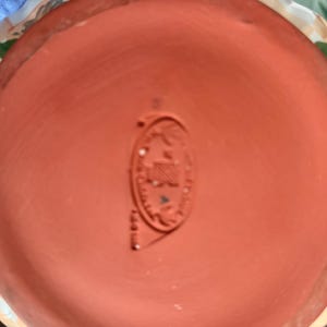 May include: A terracotta-colored ceramic pot base with a stamped emblem. The emblem is oval-shaped and features text and a design. The rim of the pot is visible, with a patterned border of white, tan, and green.