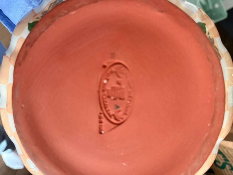 May include: A terracotta-colored ceramic pot base with a stamped emblem. The emblem is oval-shaped and features text and a design. The rim of the pot is visible, with a patterned border of white, tan, and green.