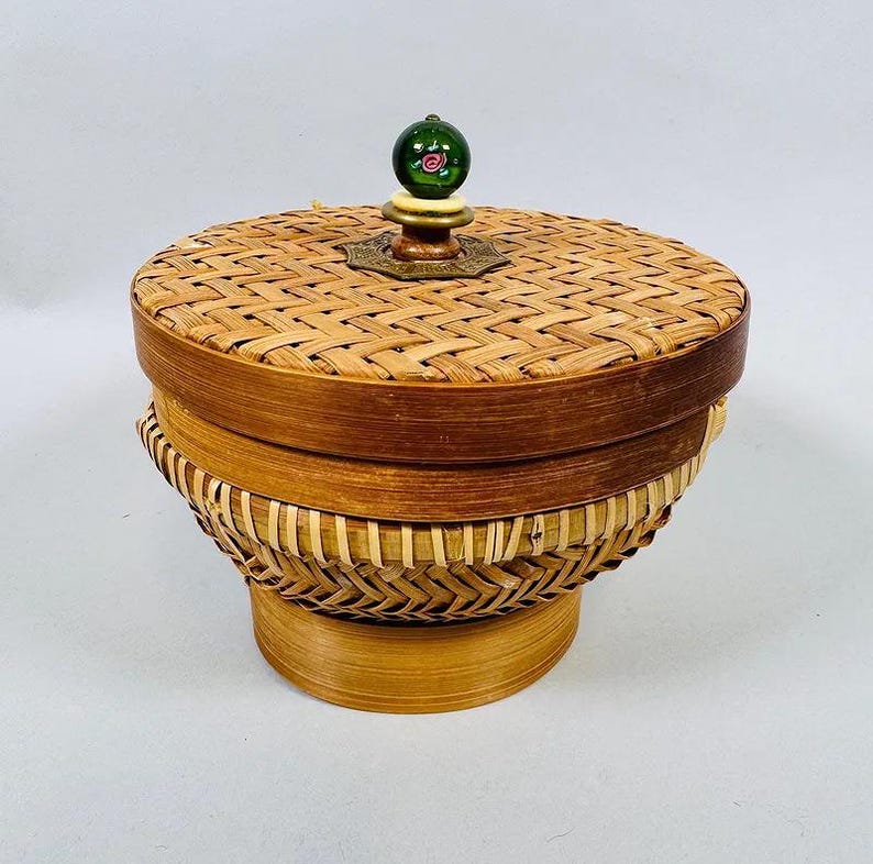 K&ouml;nnte beinhalten: Ein runder, gewebter Korb mit Deckel. Der Korb ist aus hellbraunem Korbgeflecht gefertigt und hat einen dekorativen gr&uuml;nen und goldenen Knopf oben. Der Korb hat einen Durchmesser von ca. 15 cm und ist 13 cm hoch.