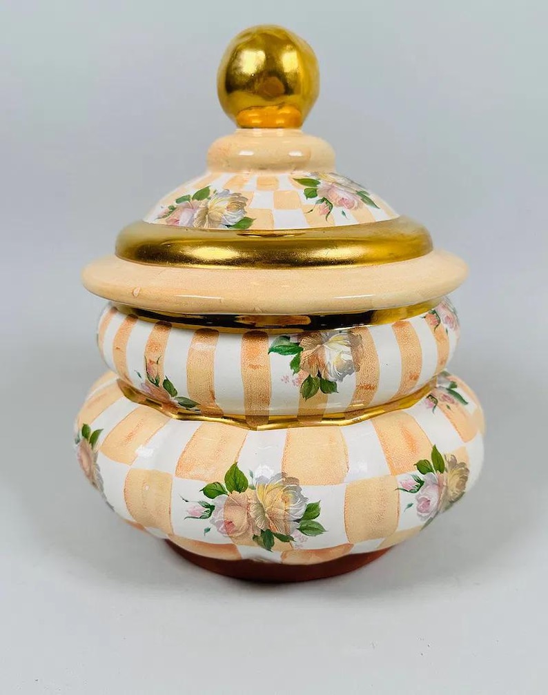 May include: A decorative ceramic jar with a lid. The jar is painted with a peach and white checkered pattern, accented with gold trim and floral designs. The lid is topped with a gold sphere.