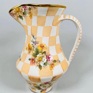 May include: A decorative ceramic pitcher with a checkered pattern in peach and white. The pitcher features floral designs and a gold rim. The handle is striped, and the pitcher is designed for serving beverages.