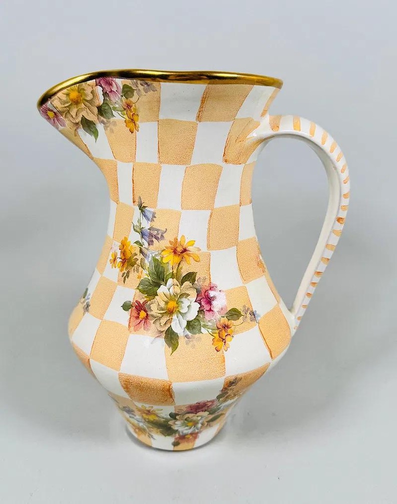 May include: A decorative ceramic pitcher with a checkered pattern in peach and white. The pitcher features floral designs and a gold rim. The handle is striped, and the pitcher is designed for serving beverages.