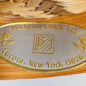 K&ouml;nnte beinhalten: Nahaufnahme einer ovalen Metallplakette mit goldenem Rand, die den Text "MacKenzie-Childs, Ltd., Aurora, New York 13026" tr&auml;gt. Die Plakette hat einen silbernen Hintergrund mit goldenen Distelmotiven und einem zentralen Logo. Die Plakette befindet sich auf einem gewebten, hellbraunen Hintergrund.