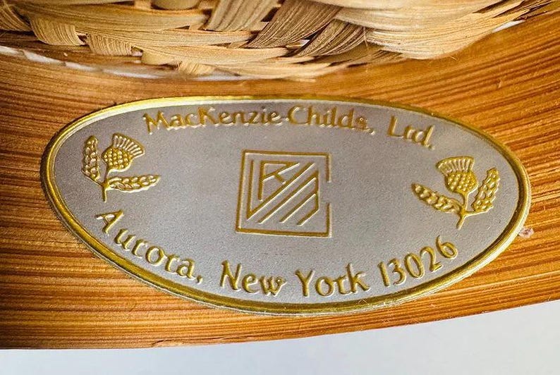 K&ouml;nnte beinhalten: Nahaufnahme einer ovalen Metallplakette mit goldenem Rand, die den Text "MacKenzie-Childs, Ltd., Aurora, New York 13026" tr&auml;gt. Die Plakette hat einen silbernen Hintergrund mit goldenen Distelmotiven und einem zentralen Logo. Die Plakette befindet sich auf einem gewebten, hellbraunen Hintergrund.