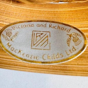 K&ouml;nnte beinhalten: Nahaufnahme eines ovalen Etiketts mit goldenem Rand, das den Text "Victoria and Richard MacKenzie Childs Ltd." tr&auml;gt. Das Etikett enth&auml;lt eine Distel und ein glockenf&ouml;rmiges Design.