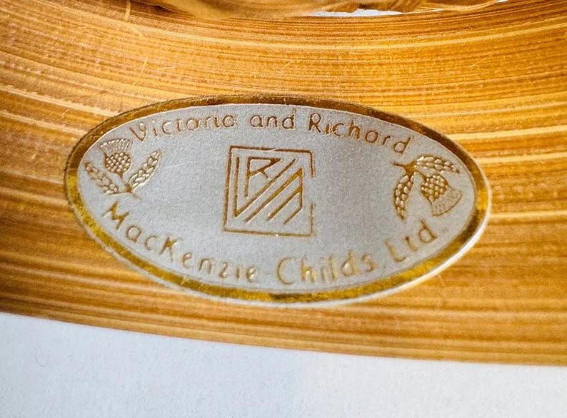K&ouml;nnte beinhalten: Nahaufnahme eines ovalen Etiketts mit goldenem Rand, das den Text "Victoria and Richard MacKenzie Childs Ltd." tr&auml;gt. Das Etikett enth&auml;lt eine Distel und ein glockenf&ouml;rmiges Design.