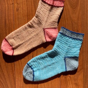 May include: Two pairs of knitted socks. One pair is tan with pink accents on the toe and heel, and a pink and red cuff. The other pair is light blue with gray accents on the toe, heel, and cuff, with a blue trim.