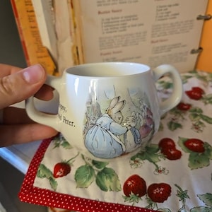 May include: A white ceramic mug with two handles, featuring a Peter Rabbit illustration in soft pastel colors. The mug sits on a strawberry-patterned cloth with a red polka-dot border. The mug has the text "Flopsy, and Peter."