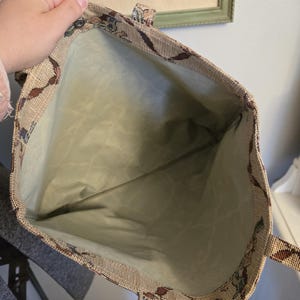 May include: Beige tapestry tote bag with a floral and leaf pattern in shades of brown, blue, and green. The interior is a light green fabric. The bag has two handles and a snap closure.