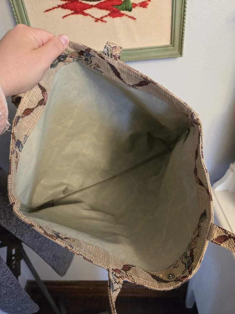 May include: Beige tapestry tote bag with a floral and leaf pattern in shades of brown, blue, and green. The interior is a light green fabric. The bag has two handles and a snap closure.