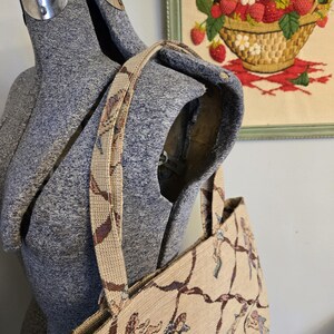 May include: Beige tapestry tote bag with a repeating pattern of cherubs and brown vines. The bag has a matching strap and is displayed on a gray dress form. A framed embroidery of strawberries is in the background.