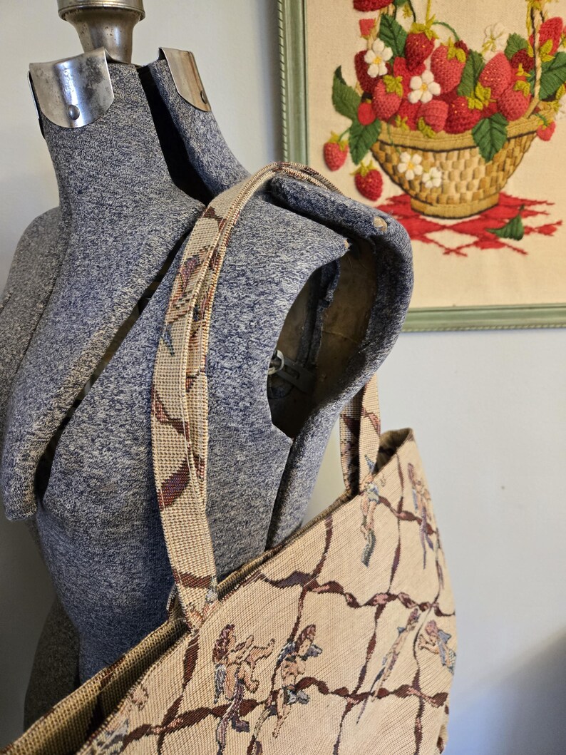 May include: Beige tapestry tote bag with a repeating pattern of cherubs and brown vines. The bag has a matching strap and is displayed on a gray dress form. A framed embroidery of strawberries is in the background.