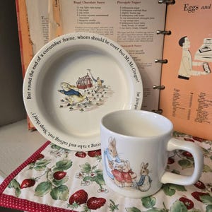 May include: A white ceramic plate and mug set featuring illustrations of rabbits and text from a children's story. The plate has a border of text. The mug and plate sit on a strawberry-patterned placemat, with a cookbook in the background.