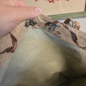 May include: A vintage tapestry tote bag with a floral pattern in shades of brown, blue, and red. The interior is a light green fabric. The bag has a snap closure and a handle.