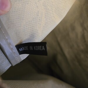 May include: Close-up of a tag inside a bag. The black tag reads "MADE IN KOREA" in white capital letters. The bag's interior is a light beige color with a textured pattern. The bag's exterior is a darker olive green.