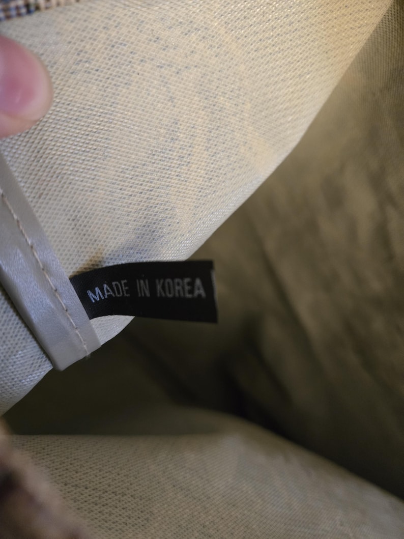 May include: Close-up of a tag inside a bag. The black tag reads "MADE IN KOREA" in white capital letters. The bag's interior is a light beige color with a textured pattern. The bag's exterior is a darker olive green.