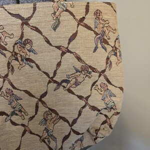May include: Beige tote bag with a repeating pattern of cherubs and brown ribbon-like lines. The cherubs have blue, purple, and pink accents. The bag has a matching handle.