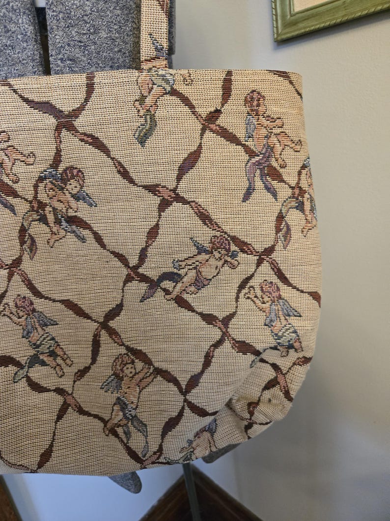 May include: Beige tote bag with a repeating pattern of cherubs and brown ribbon-like lines. The cherubs have blue, purple, and pink accents. The bag has a matching handle.