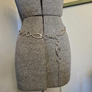 May include: Silver-toned chain belt with oval and circular links, draped on a gray dress form. The belt features a dangling chain with a large oval pendant and small rings at the end. The dress form is on a metal stand.