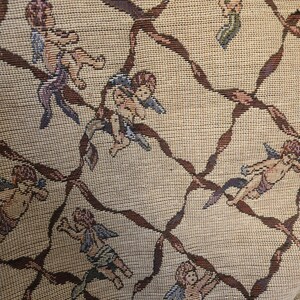 May include: Beige fabric with a repeating pattern of cherubs and brown ribbon-like lattice. The cherubs are depicted in various poses, with wings and draped clothing in shades of blue, purple, and green. The fabric has a woven texture.