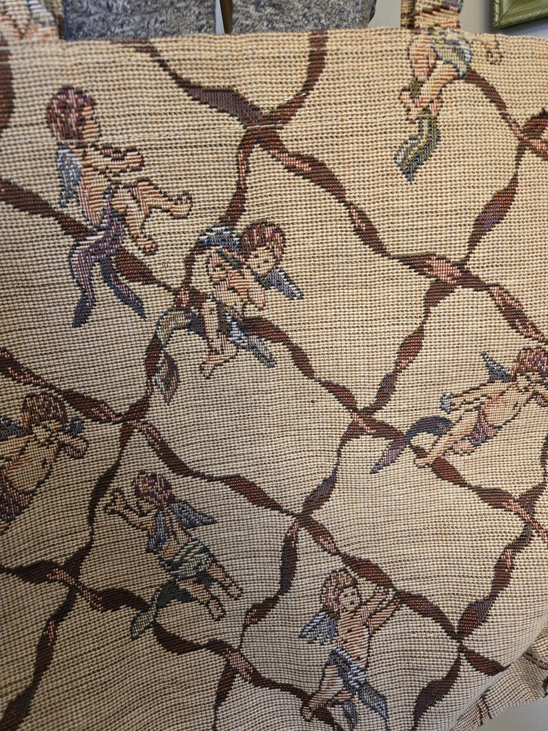 May include: Beige fabric with a repeating pattern of cherubs and brown ribbon-like lattice. The cherubs are depicted in various poses, with wings and draped clothing in shades of blue, purple, and green. The fabric has a woven texture.
